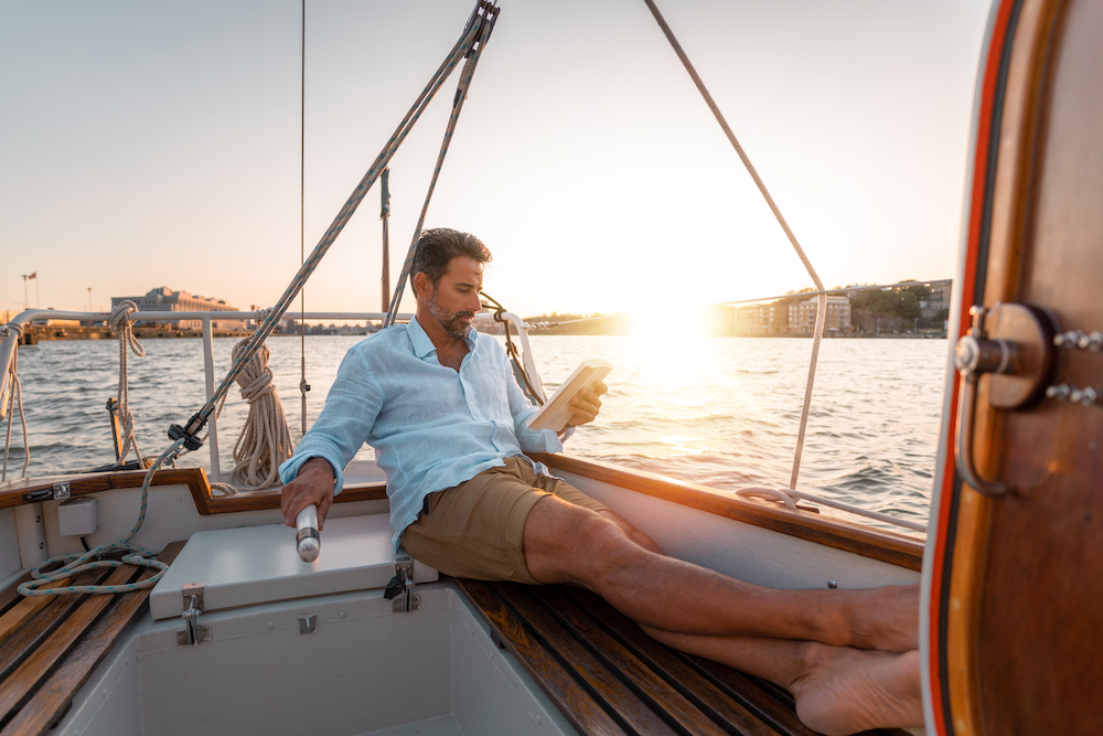 A man reading a book on a sail boat as the sun is setting
