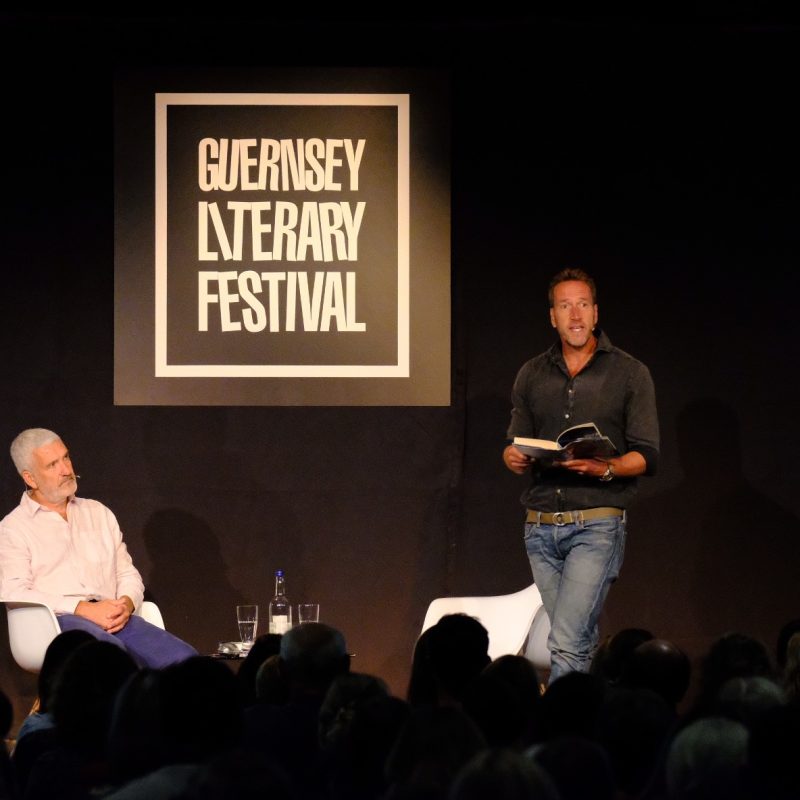A man with white hair sitting down on stage listening to another gentleman reading a section from a book at the Guernsey Literary Festival