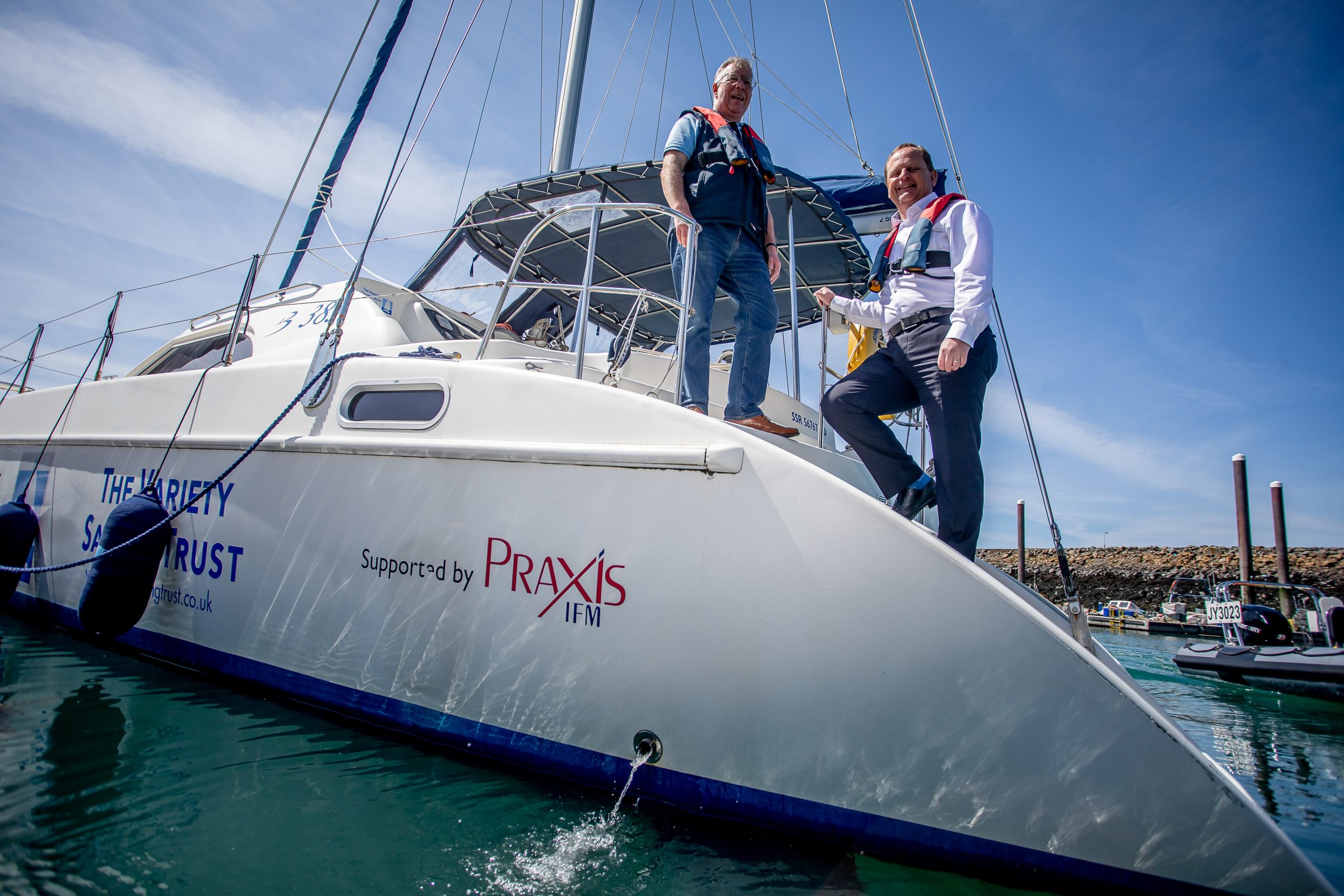 Two men stood on a sailing boat with life jackets on. The boat is in the water and has the PraxisIFM logo printed on it